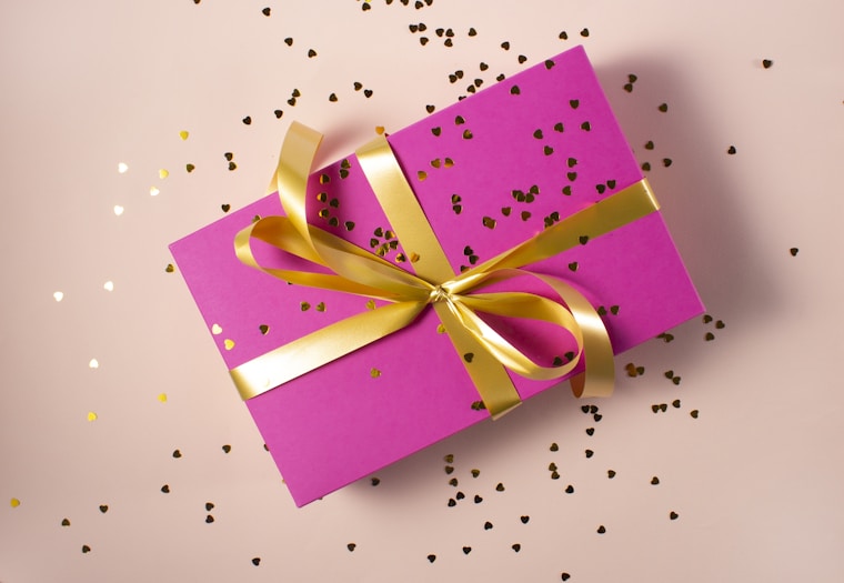 A pink gift box with a gold ribbon is surrounded by small gold heart-shaped confetti on a light pink surface.