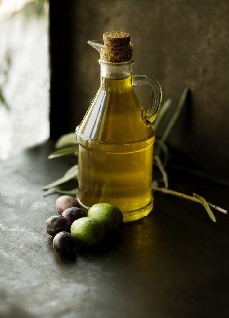 A glass bottle of olive oil with a cork stopper is placed next to several green and purple olives and olive branches on a dark surface.