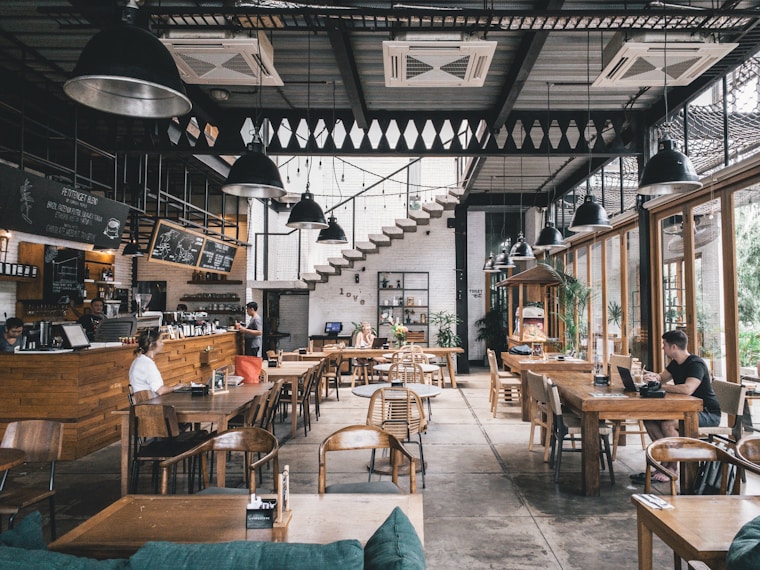 Spacious modern café with wooden tables and chairs, large windows, indoor plants, and several people working or relaxing. Counter and menu visible on the left, staircase in the background.