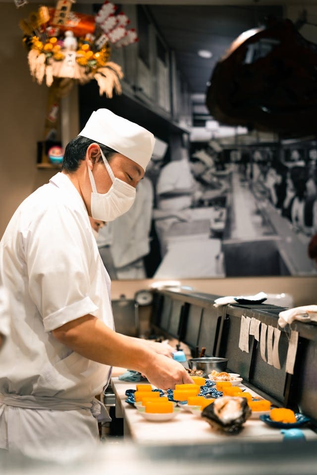 A sushi chef in a white uniform and face mask prepares food at a counter, with various dishes in front of him and a black-and-white kitchen photo on the wall behind.