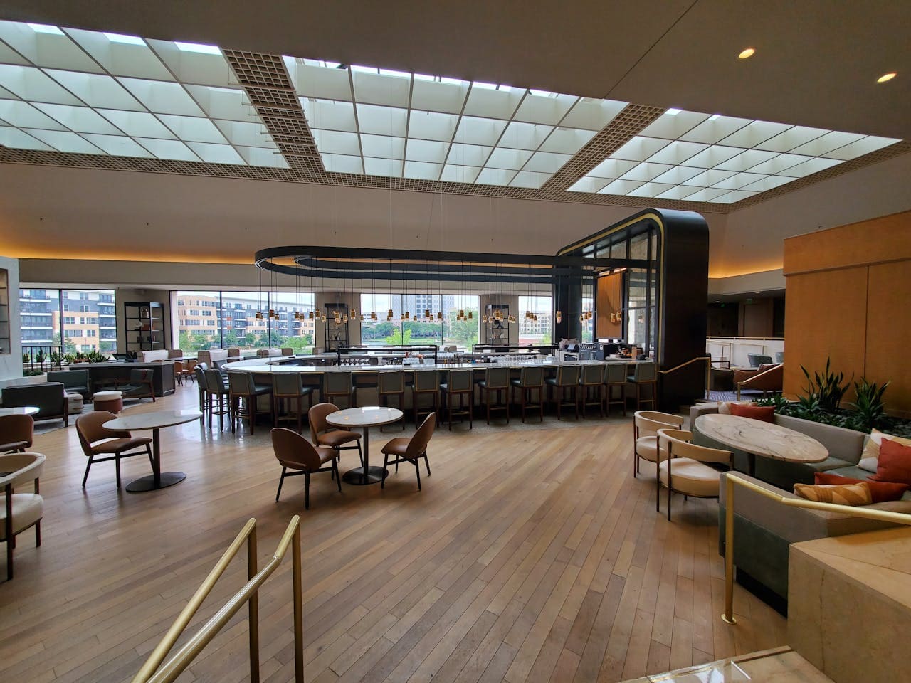 Spacious modern hotel lobby with wooden floors, tables, chairs, a large central bar, and floor-to-ceiling windows providing natural light.