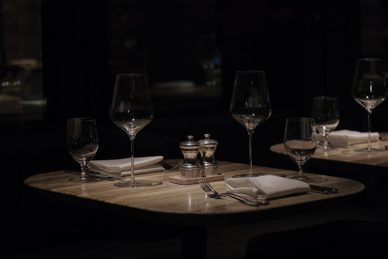 A dimly lit restaurant table set with empty wine glasses, water glasses, white napkins, silverware, and salt and pepper shakers.